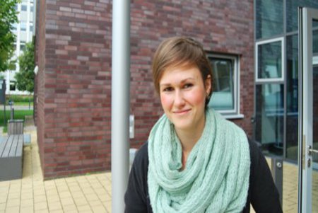 Ein Frau mit kurzen Haaren und türkisem Schal, schaut vor einem Gebäude der FH Kiel in die Kamera.