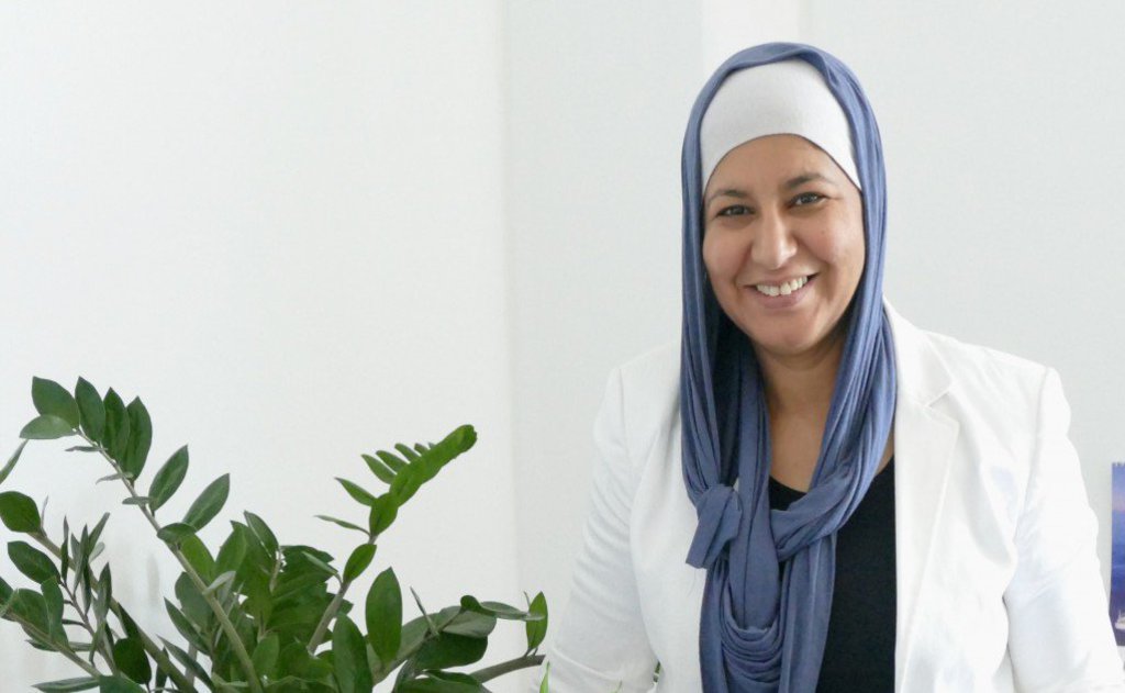 Eine Frau in weißem Jacket lächelt in die Kamera. Sie trägt zudem ein locker sitzendes Kopftuch.