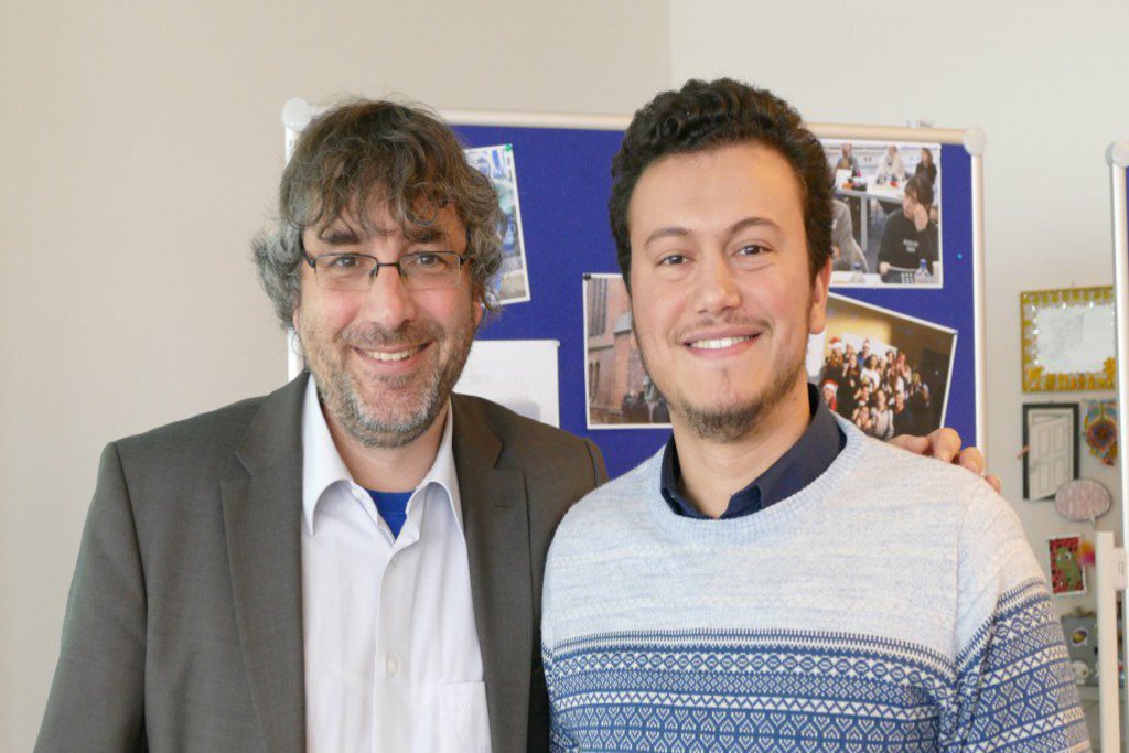 Ein älterer und ein jüngerer Mann posieren vor einer Infowand.