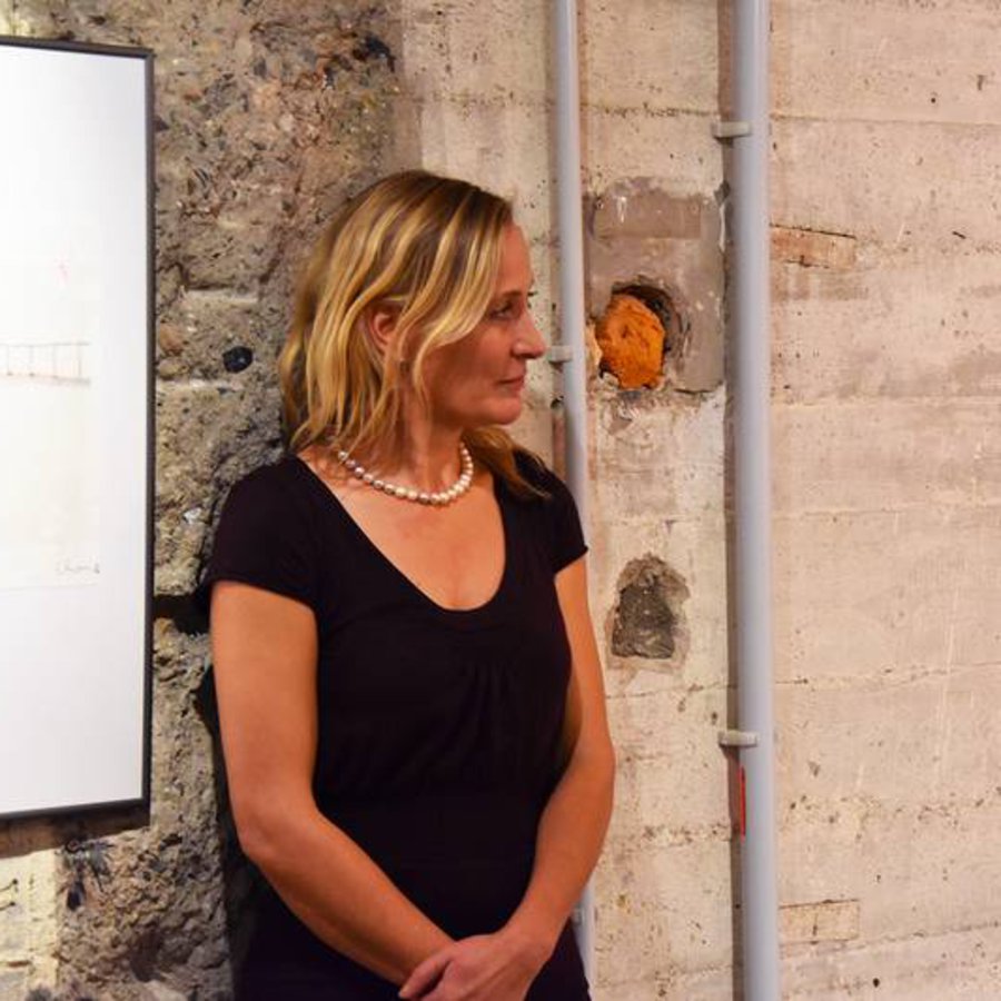 Eine Frau in blauer Bluse, steht vor einer Rustikalen Wand und verschränkt die Arme.