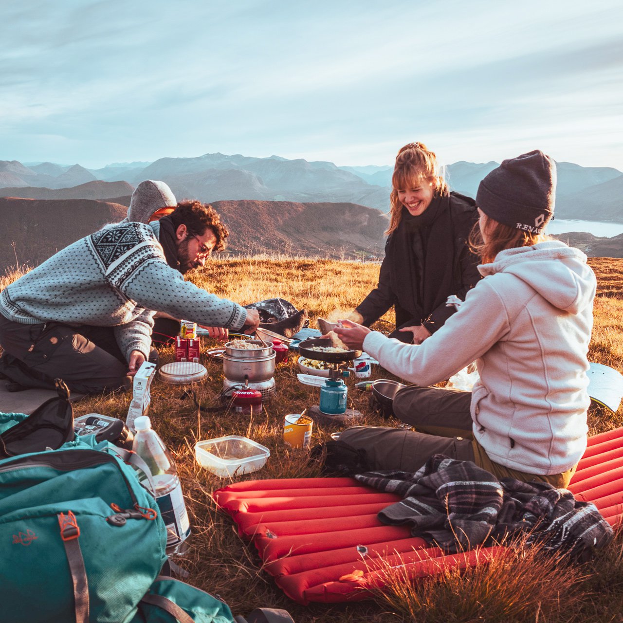 Junge Menschen sitzen mit Wanderausrüstung auf einem Bergplateau und bereiten Essen auf Campingkochern zu.