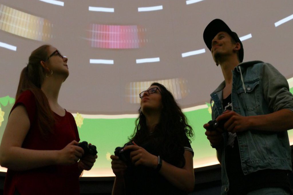 Zwei Frauen und ein Mann sind mit Controllern ausgestattet und schauen auf eine bespielte Kuppel.