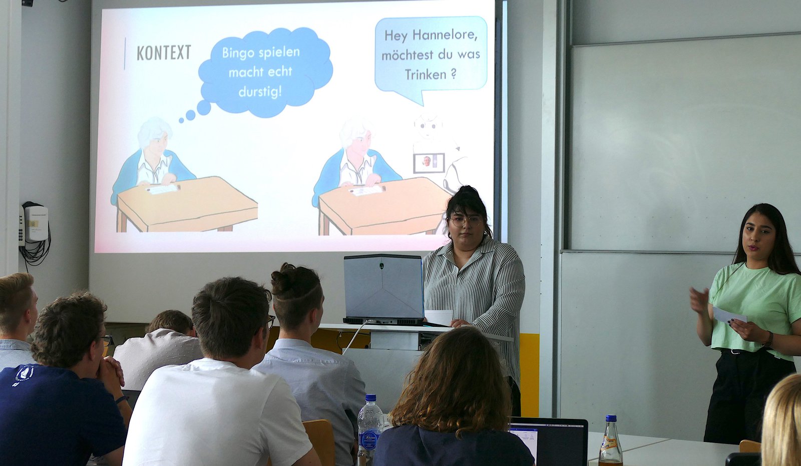 Ein Hörsaal gefüllt mit Studierenden. Zwei Studentinnen halten einen Vortrag zum Service-Roboter.