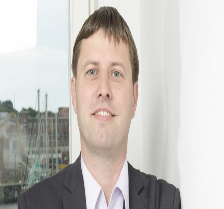 Ein Mann in hellem Hemd und schwarzem Sakko verschränkt die Arme und lächelt freundlich in die Kamera.