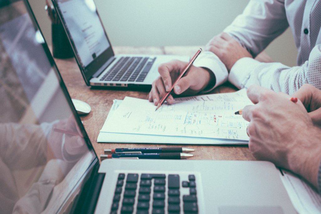 Zwei Laptops stehen auf einem Holztisch, zwischen ihnen liegen Notizzettel auf denen Hände am werkeln sind.