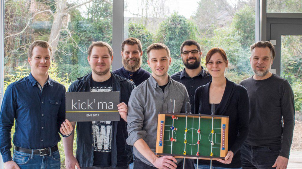 Eine Gruppe posiert vor einer Fensterfront und hält dabei ein Schild und einen Mini-Kicker-Tisch in den Händen.