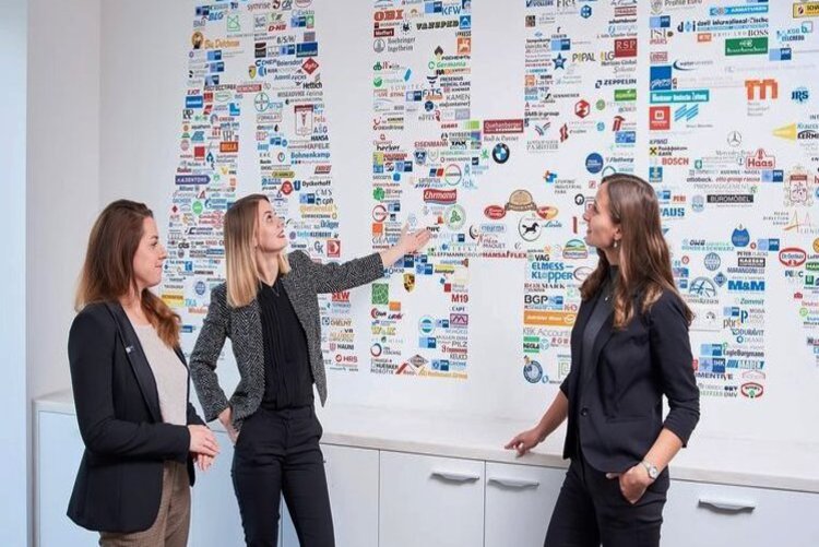 Drei Frauen posieren vor einer Wand, die mit den Buchstaben AHK versehen ist. Die Buchsataben sind sehr groß, bunt und bestehen aus vielen kleinen Markenlogos.