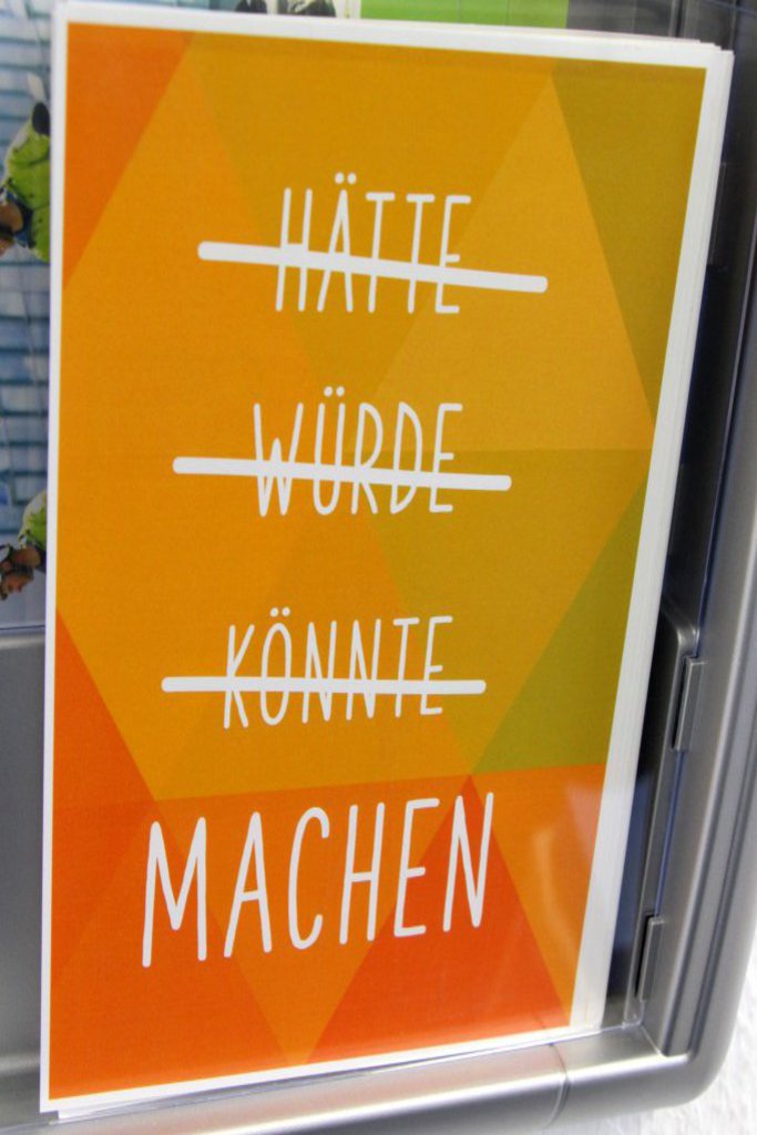 Auf dem Plakat sind drei durchgestrichene Worte zu sehen: "HÄTTE", "WÜRDE", "KÖNNTE" und darunter das nicht durchgestrichene Wort: "MACHEN".