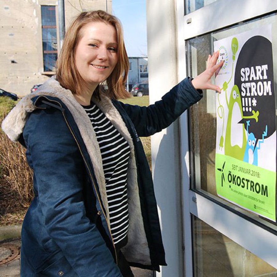 Eine Frau ist gerade dabei ein Plakat anzubringen und lächelt dabei motiviert in die Kamera.