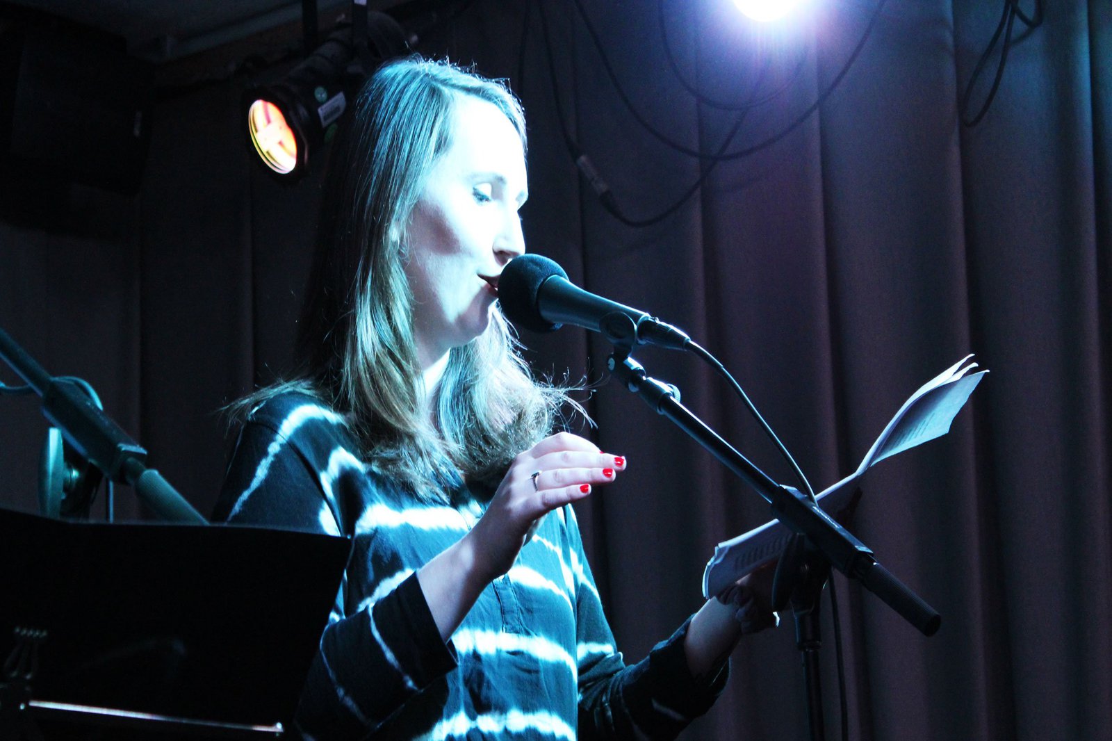 Eine Frau steht vor einem Mikrofon, auf einer schwach beleuchteten Bühne und liest etwas aus dem Skript in ihrer Hand ab.