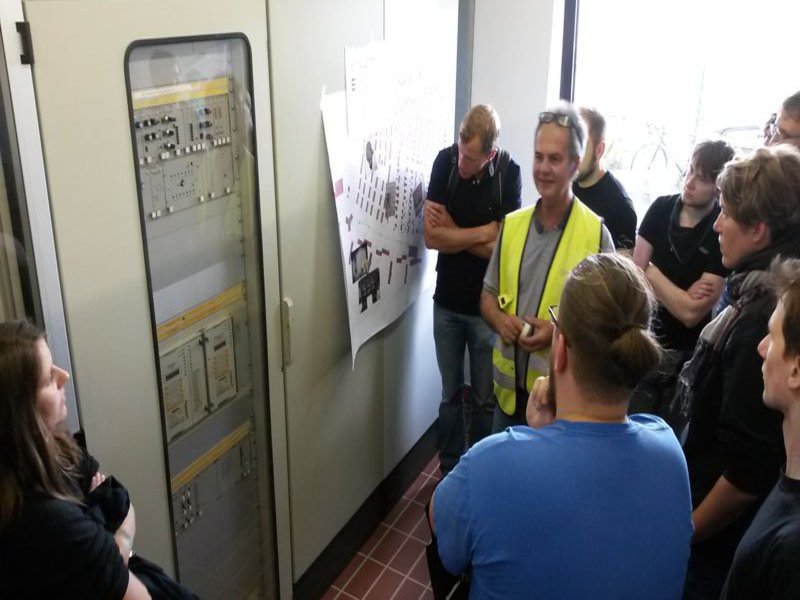 Ein Mann in gelber Warnweste zeigt einer Gruppe Studierender einen Steuerungskasten.