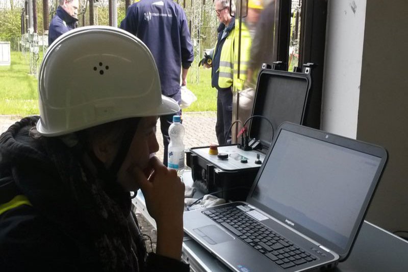 Eine Frau mit Helm auf dem Kopf schaut auf einen Laptop. Im Hintergrund sind drei Männer und besprechen etwas.