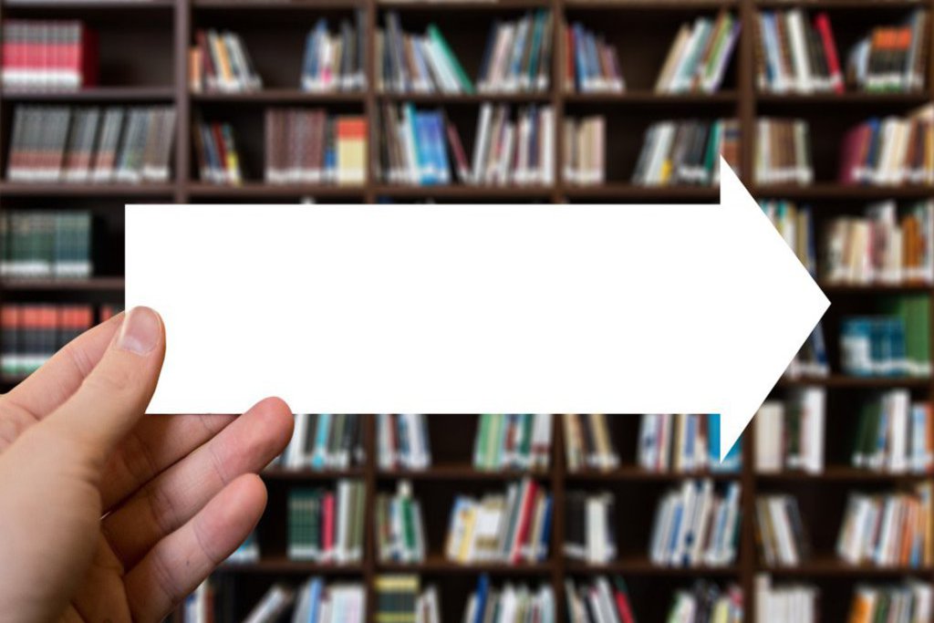 Eine Hand hält einen Papierpfeil vor ein gefülltes Bücherregal.