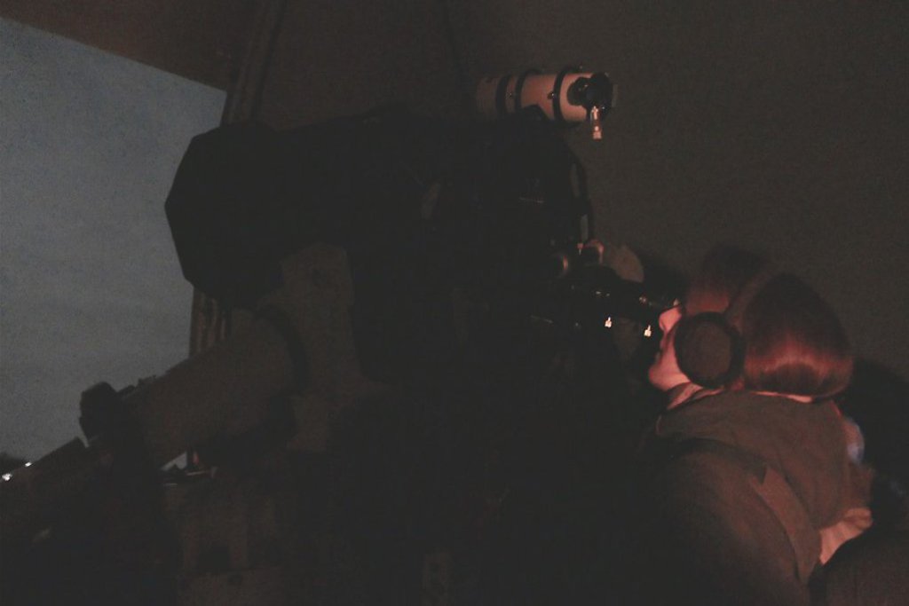 Eine FRau mit Ohrenschützern schaut durch ein Teleskop in den Abendhimmel hinaus.