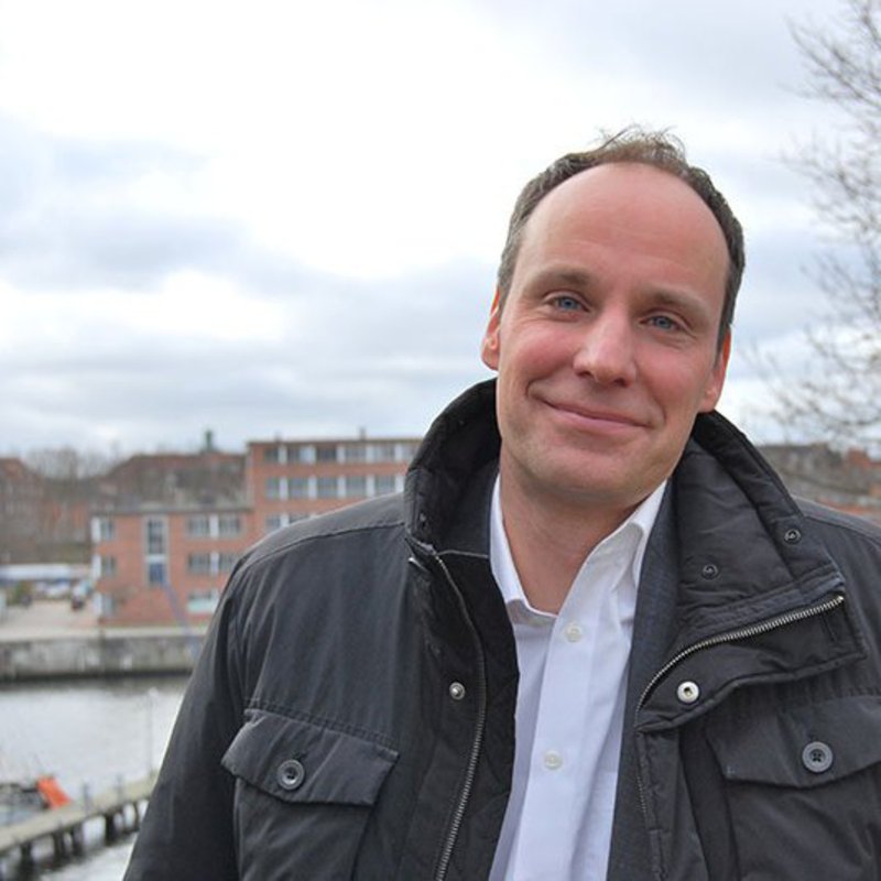 Ein Mann in Winterjacke und weißem Hemd, schaut Nahe der Schwentine freundlich in die Kamera.