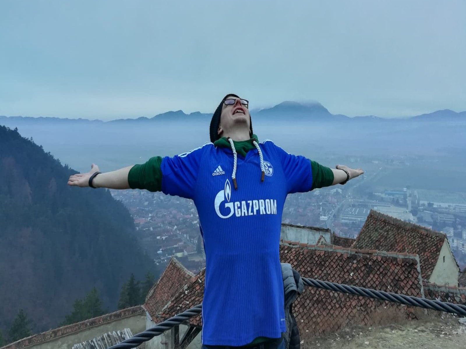 Ein Mann in einem Schalke 04 Trikot posiert mit weit geöffneten Armen auf einem Dach. Im Hintergrund ist eine von Bergen umgebene und in Nebel getauchte Stadt.