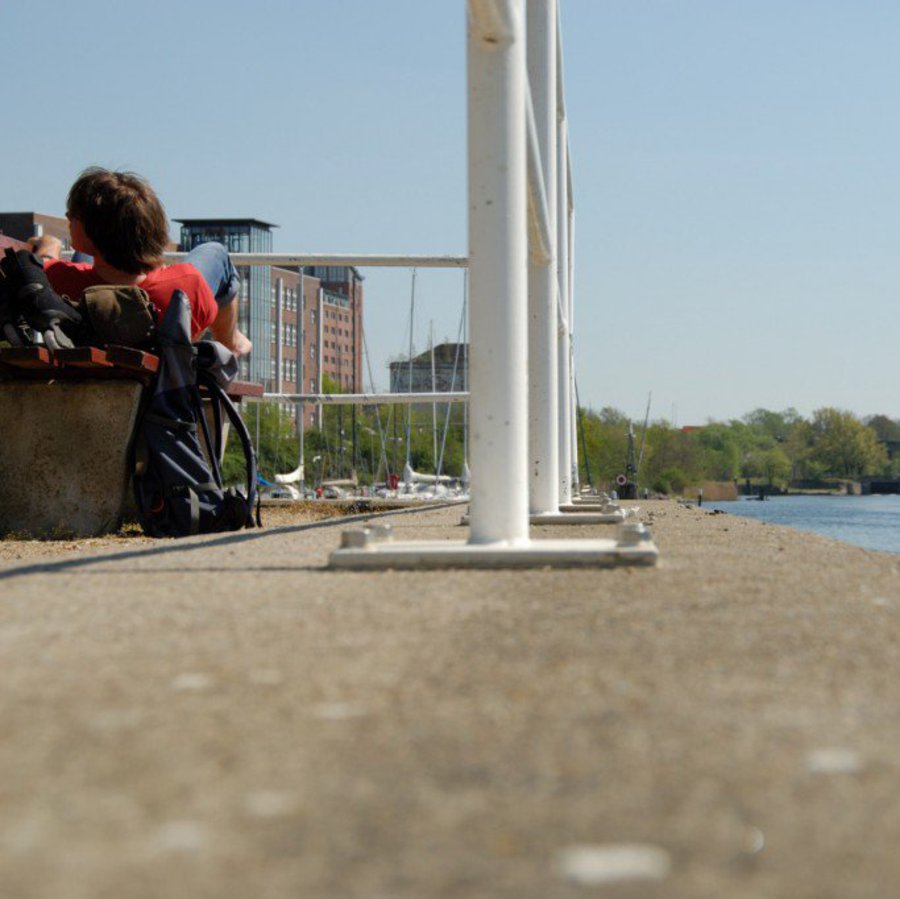 Im Hintergrund liegt eine Person bei bestem Wetter auf einer Bank. Davor sieht vom Boden aufgenommene, in den Boden eingelassene Metall Stangen. Rechts ist das Meer zu sehen.