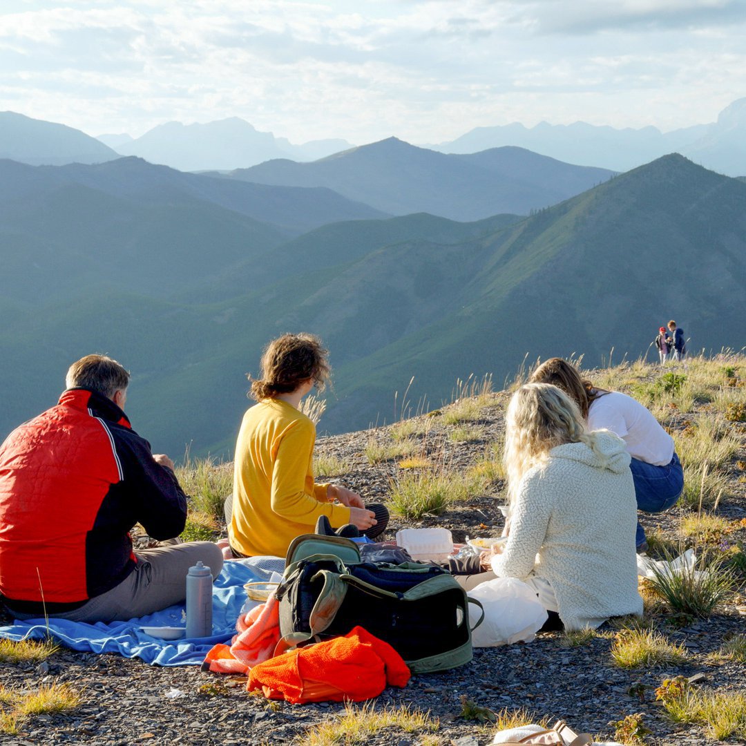 Junge Menschen sitzen beim Picknick auf einem Bergplateau und blicken ins Tal