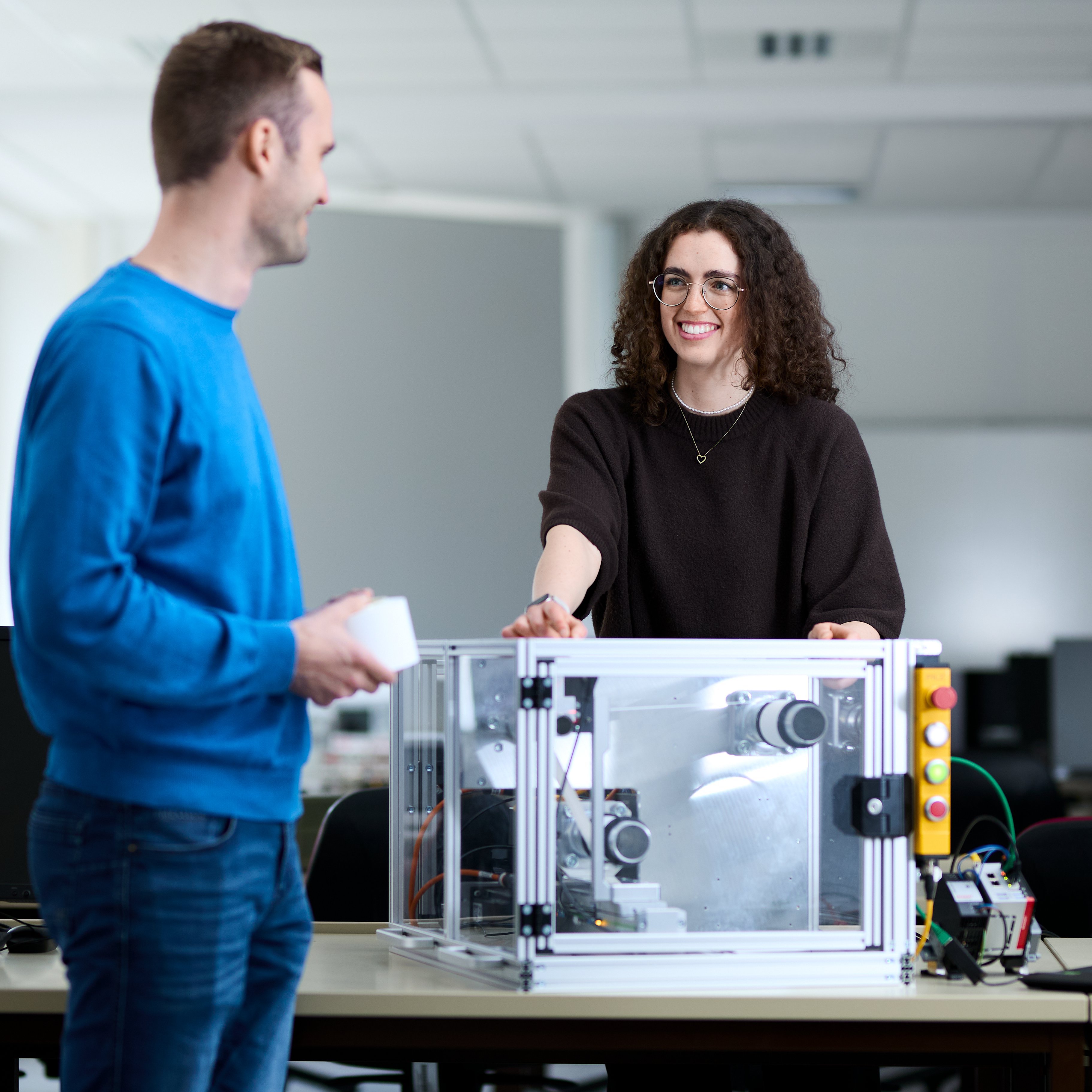 Eine junge Frau steht hinter einem Tisch in einem Labor, vor dme Tisch steht ein junger Mann, er ist von hinten zu sehen