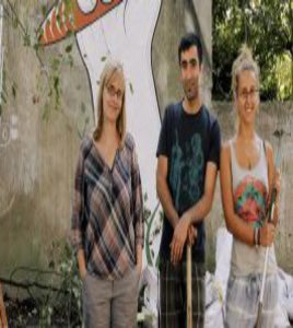 Ein Mann und zwei Frauen stehen vor einem Grafitti, dass sich in einem Garten befindet.