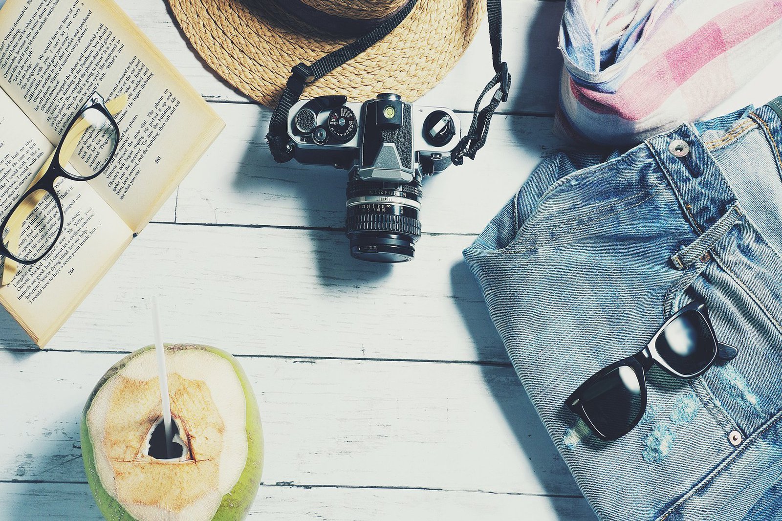 Eine Trinkkokosnuss, ein Buch und eine Brille, ein Hut, eine Kamera, ein Schal, eine Jeans und eine Sonnenbrille liegen auf einer Picknickbank.