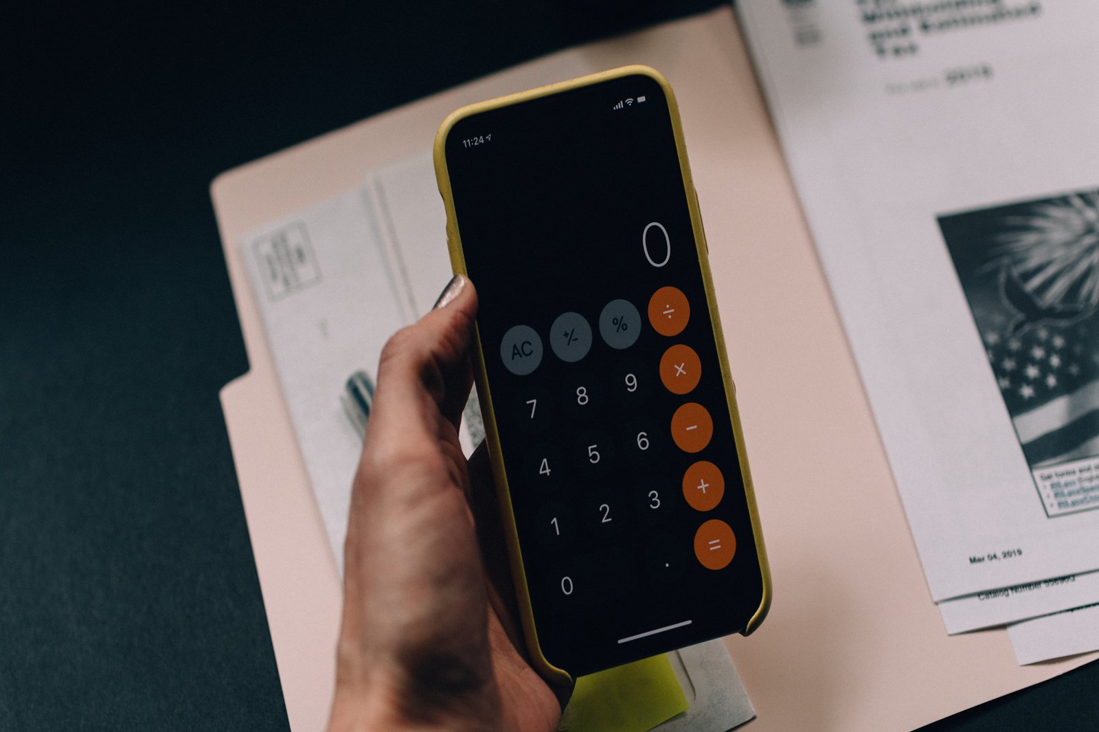 Ein Handy mit geöffneter Taschenrechner-App, gehalten in einer weiblichen Hand. Im Hintergrund sind Papiere zu sehen.