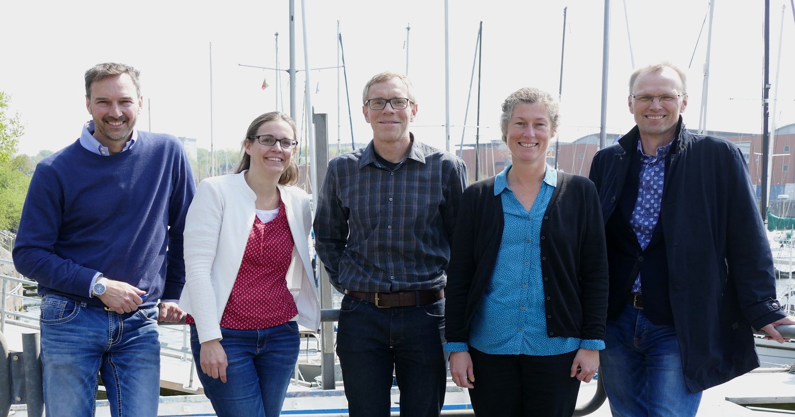 Zwei Frauen und drei Männer posieren vor dem Hafen der Schwentinemündung und lächeln in die Kamera.