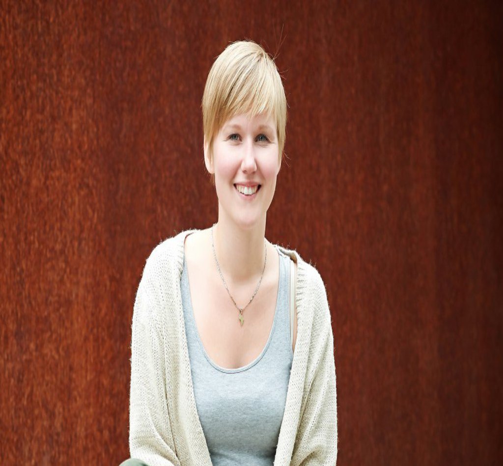 Eine Frau in grauem Top und grüner Hose, sitzt im Schneidersitz auf dem Boden und schaut freundlich in die Kamera.