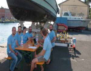 Eine Gruppe in hell blauen Shirts, sitzt an ein paar Bierbänken und einem dazugehörigen Holztisch.