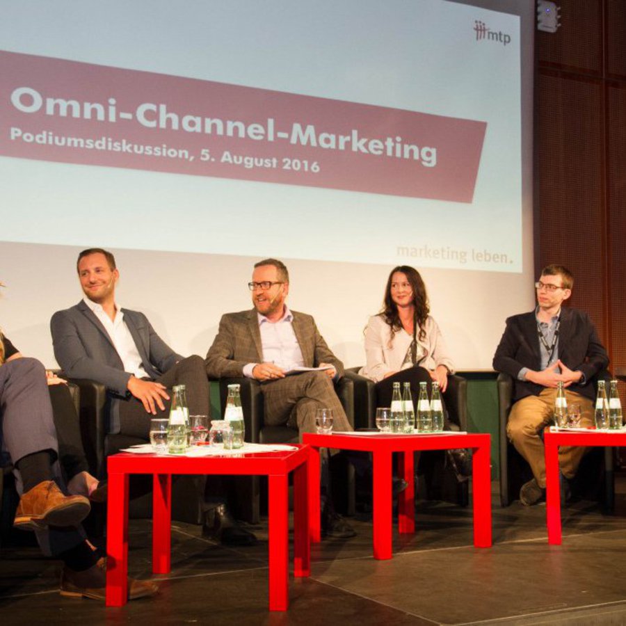 Sieben Personen sitzen auf einer Bühne in einer Diskussionsrunde. Auf drei roten BEistelltischen stehen Wasserflaschen.