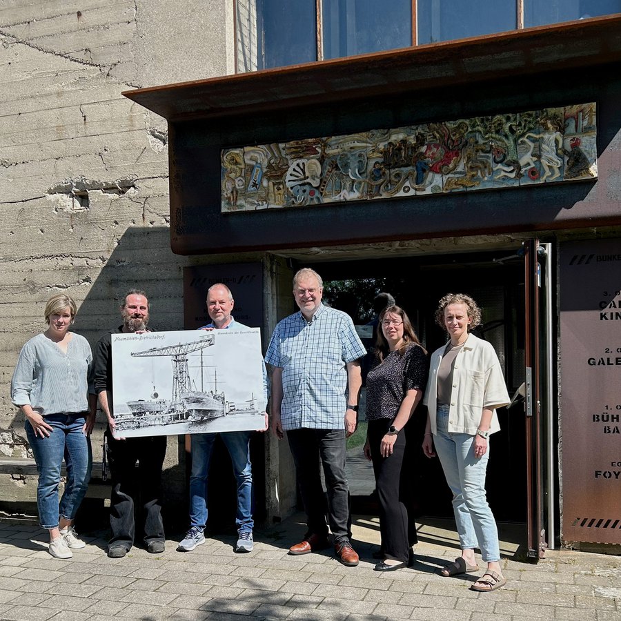 Vor dem Eingnag des Kulturzentrums BUnker-D der FH Kiel steht das Organisationsteam und zeigt die Bildtafel mit dem Herzstück der Ausstellung. „Oma“ lautete der Spitzname des einstigen Riesenkrans der Howaldtswerke. (v.l.n.r.) Kristiina Thiel und Jan Pieper (ZKW), Wolfgang D. Kuessner (Sammler), Markus Schack und Claudia Tippmann (ZKW), Anja Konzan (Büro Soziale Stadt Neumühlen-Dietrichsdorf). 