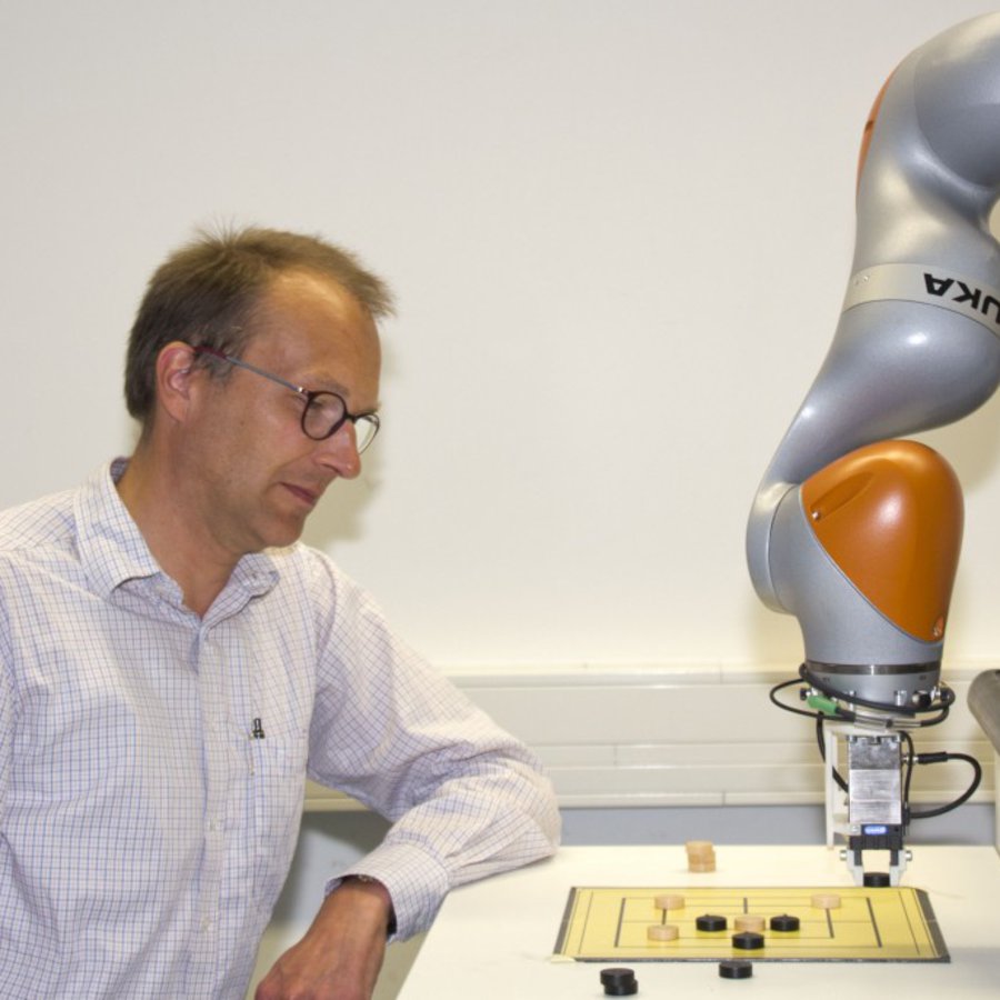 Ein Mann in wieß-kariertem Hemd spielt Mühle gegen einen Roboter.