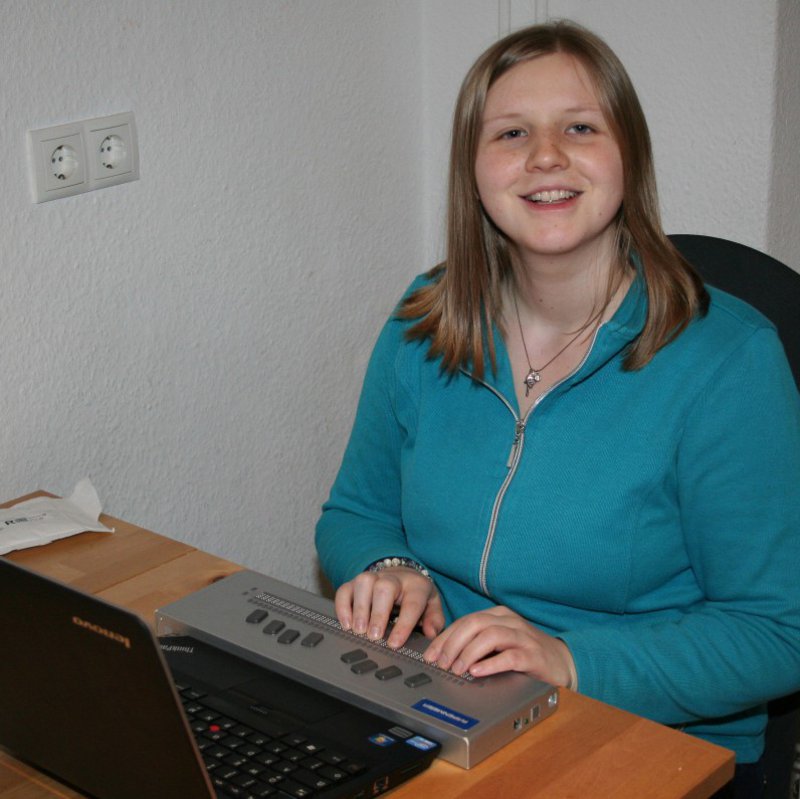 Eine Frau in grünem Pullover sitzt vor ihrem Laptop und berührt mit den Händen, ein davor geschaltetes Gerät.