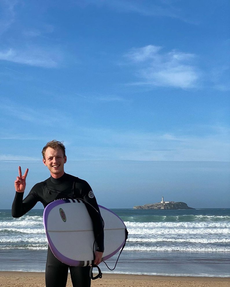 Tim lächelt und winkt in die kamera, er trägt einen Neoprenanzug und ein Surfbrett unter dem Arm