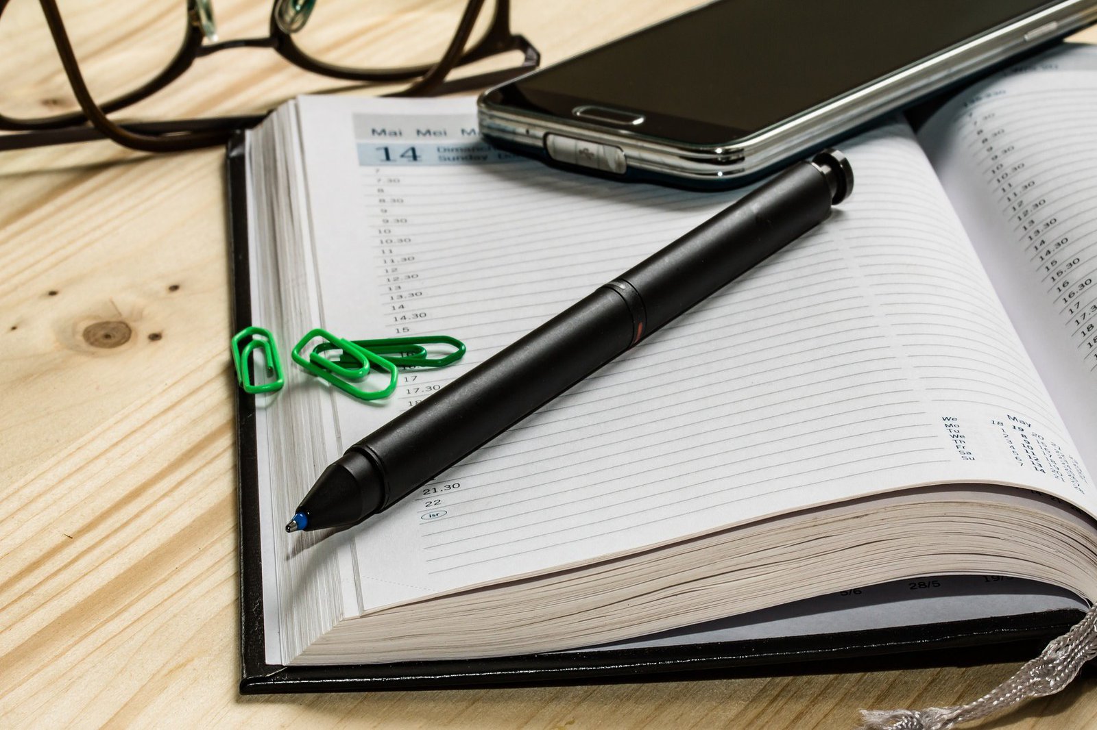 Kalender, Büroklammern, Kugelschreiber, Handy und Brille liegen verteilt auf einem Holztisch.