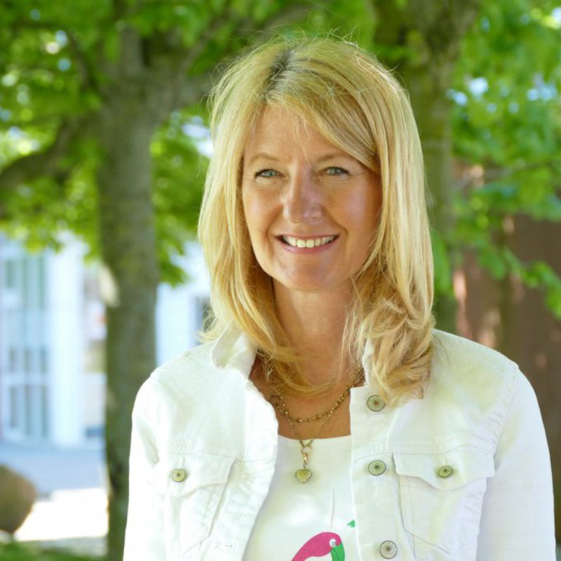 Eine Frau in weißer Sommerjacke steht im freien und lächelt in die Kamera.