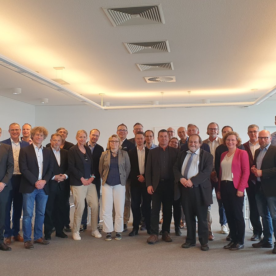 Auf dem Bild sind 31 Personen zu sehen, die als Gruppe im Halbkreis stehen. Sie sind business casual gekleidet und lächeln in die Kamera. 