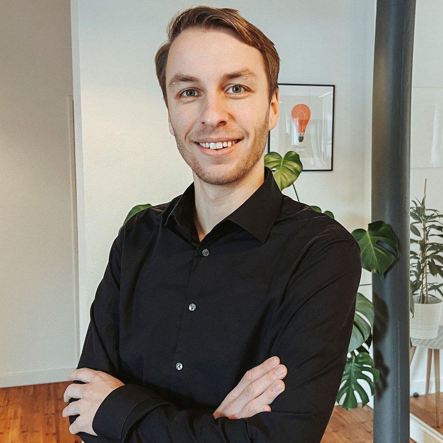 Ein junger Mann in dunkelblauem Hemd posiert mit verschränkten Armen und lächelnd.