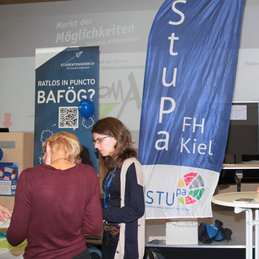 2 Studentinnen und 2 Studenten stehen in einem größeren Raum an Stehtischen, es ist eine Beachflag mit der Aufschrift StuPa zu sehen und ein Aufsteller vom Studentenwerk, die für Bafög-Beratung wirbt.