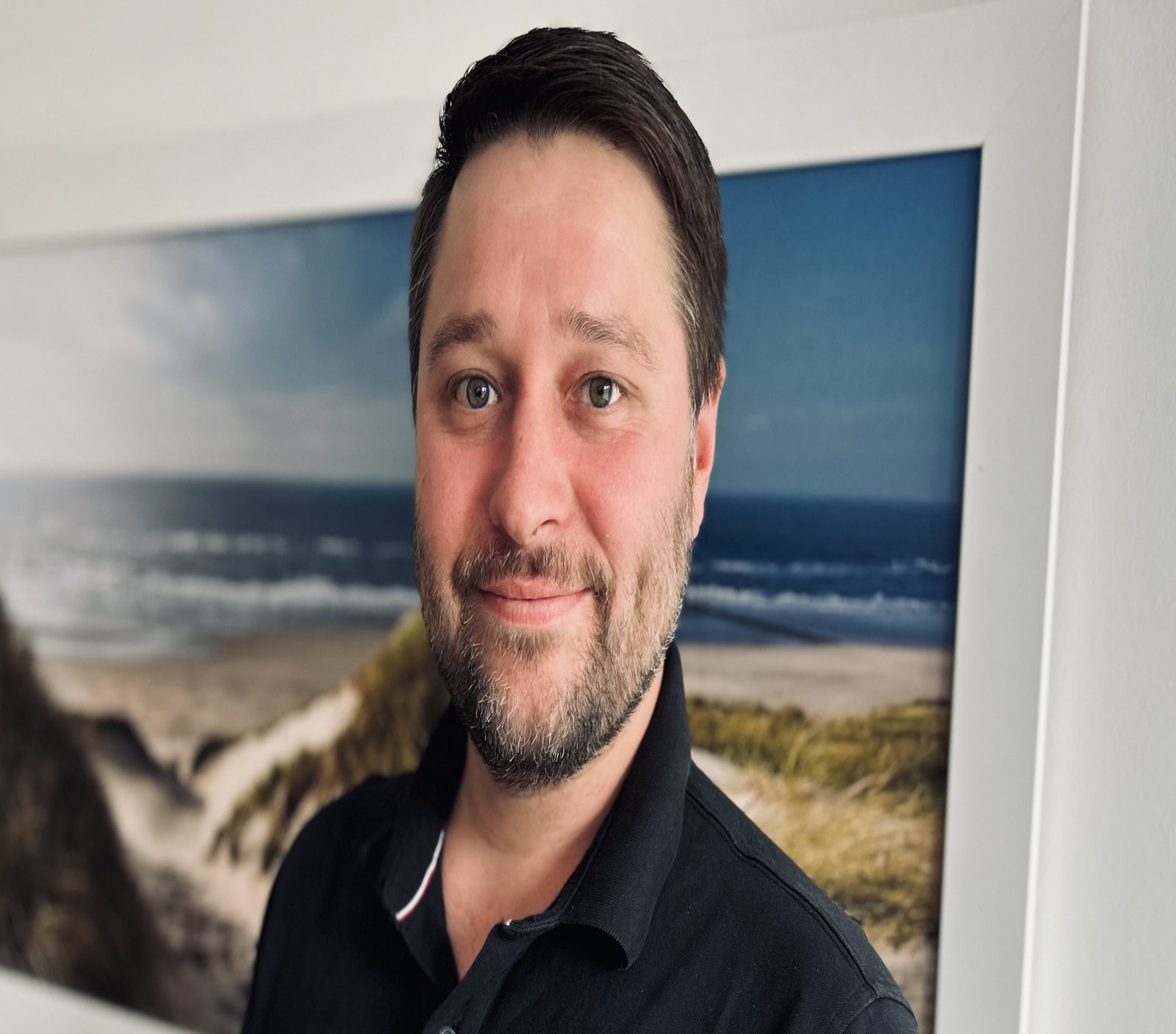 a bearded man standing in front of a picture showing a coastline