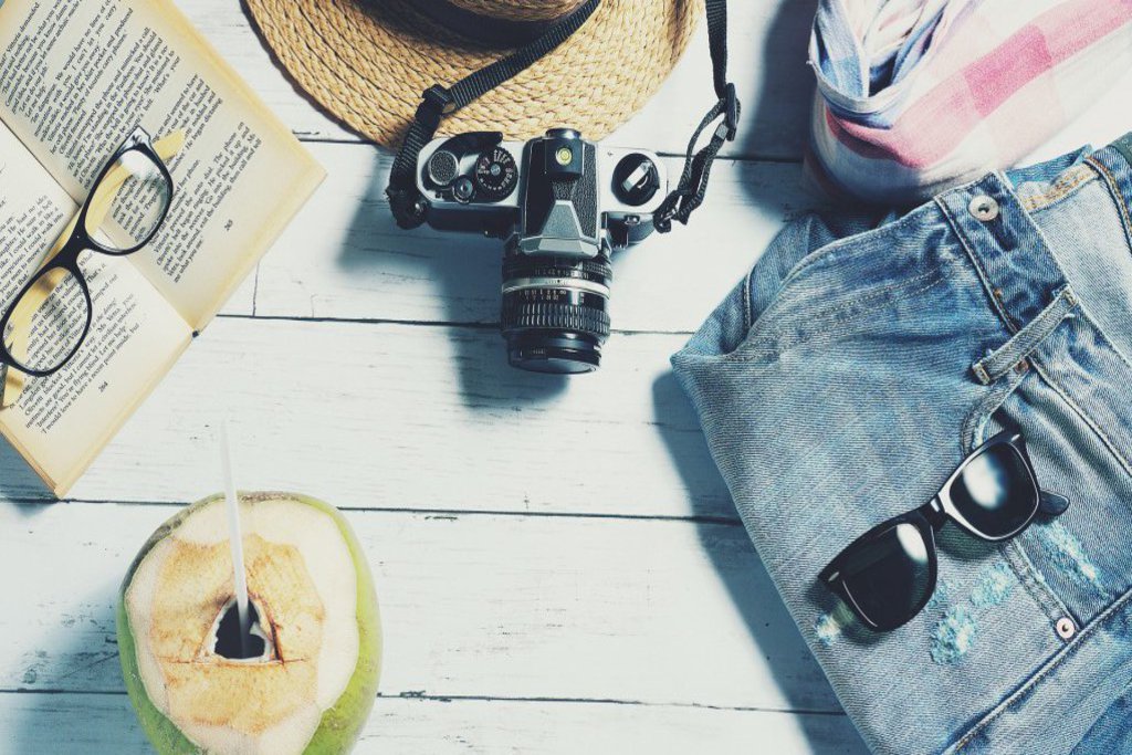 Eine Kokosnuss, ein Buch mit Brille darauf, ein Hut, eine Kamera, ein Schal und eine Jeans mit Sonnenbrille darauf liegen auf einem Holztisch.