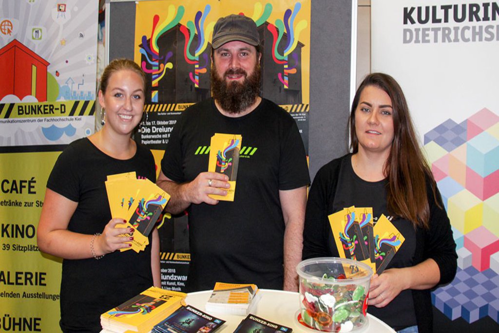 Zwei Frauen und ein Mann präsentieren Flyer und posieren vor einer Plakatwand. Die Farbe Gelb sticht in dem Bild deutlich hervor.