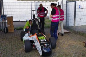 Unter einem Pavillon steht ein bemannter Rennwagen und neben ihm zwei Crew-Mitglieder.