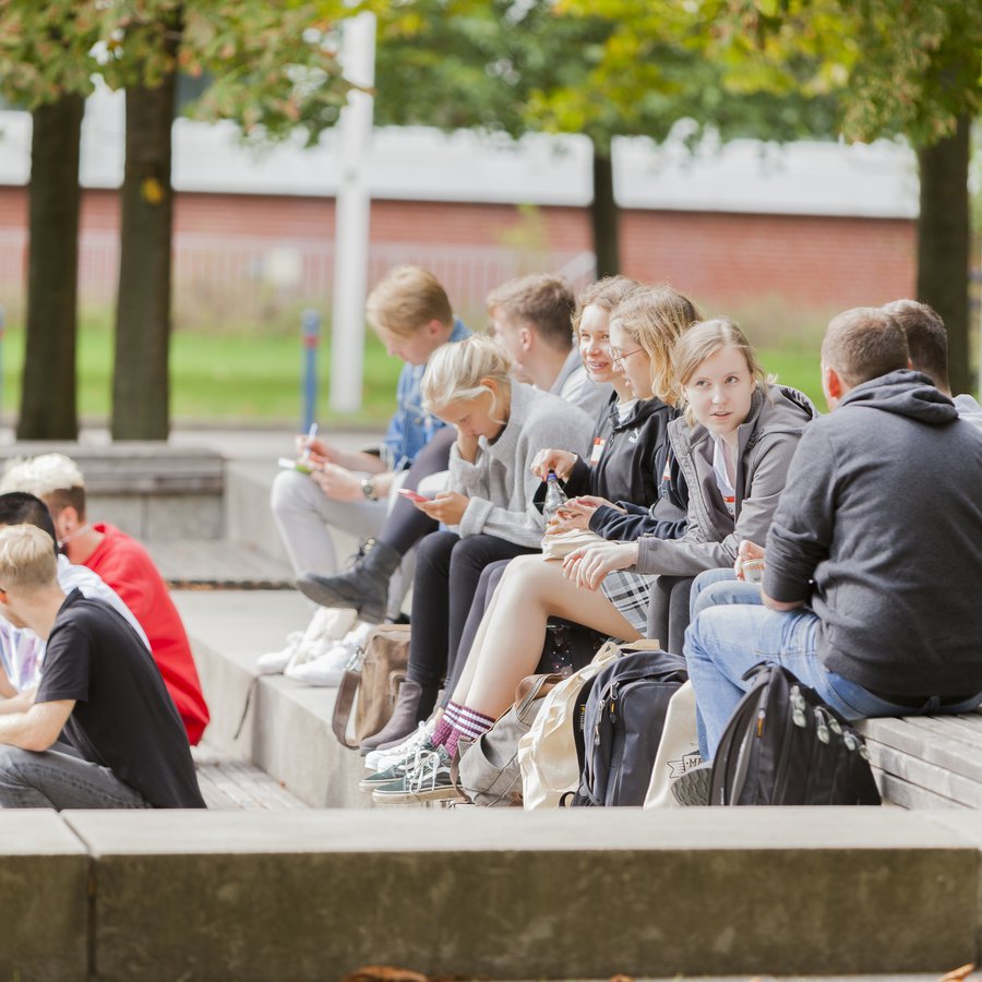Campusimpression Eine Gruppe Studierender sitzt auf dem Campus, die Sonne scheint.