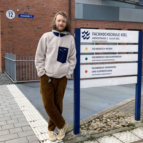 Ein junger Mann steht an ein Hinweisschild gelehnt und schaut lächelnd in die Kamera. Auf dem Schild steht: "Fachhochschule Kiel", "Fachbereich Medien", "Fachbereich Maschinenwesen", "Fachbereich Informatik und Elektrotechnik".