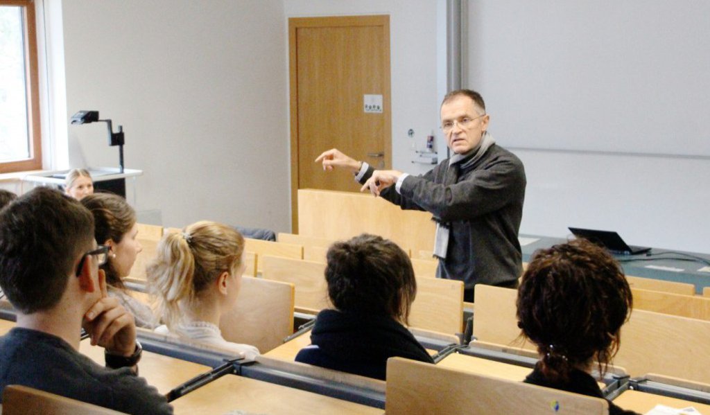 Ein Professor steht an der ersten Stuhlreihe eines Hörsaals und spricht zu den dahinter sitzenden Studierenden.