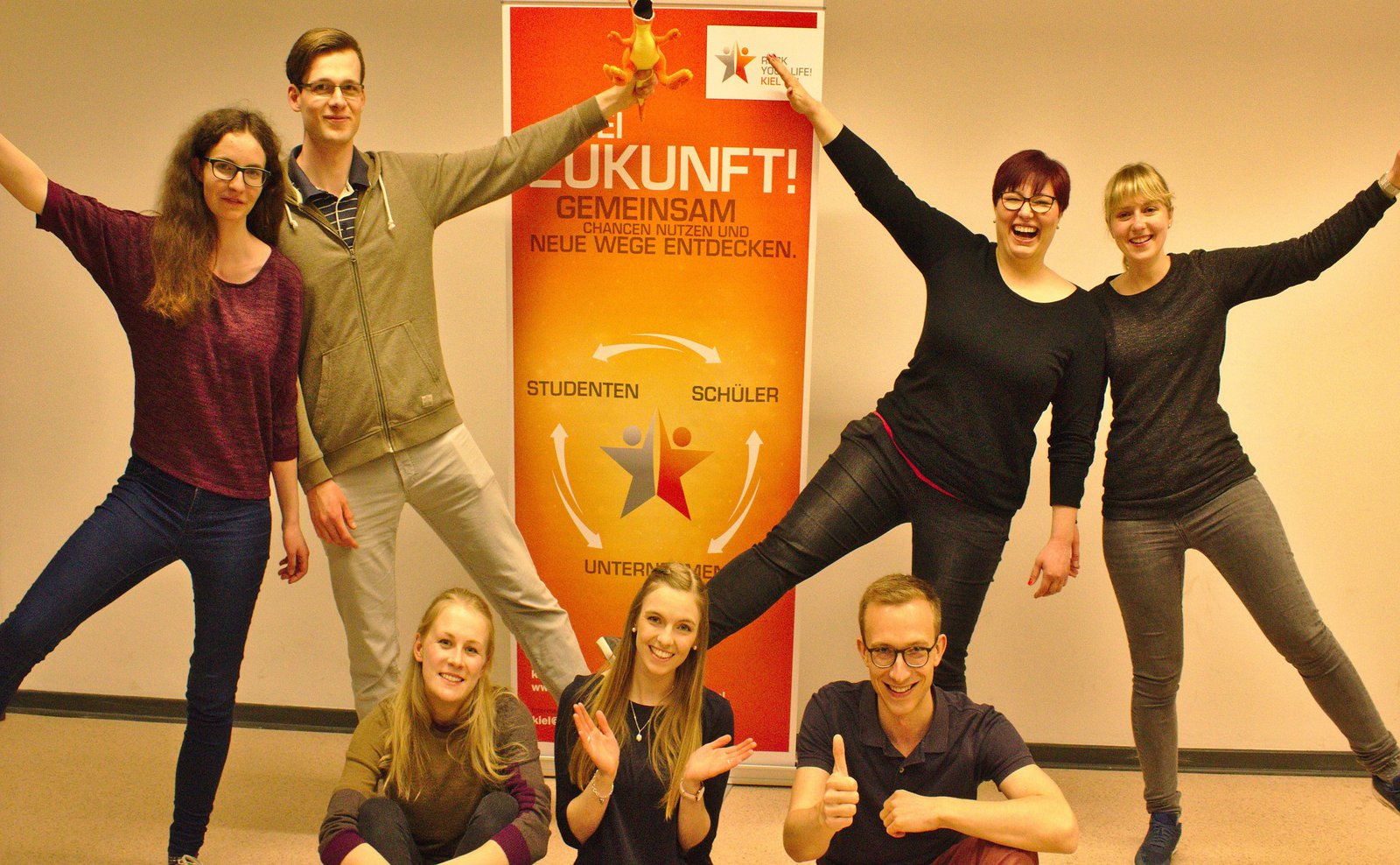 Studierende posieren freudig vor einem Plakat.