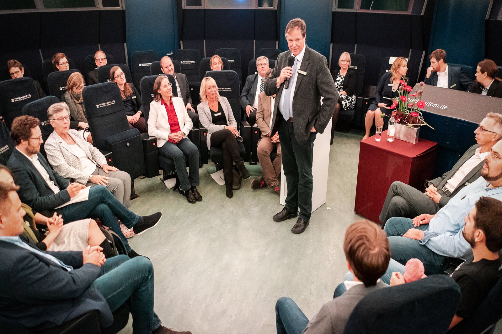 Eine GRuppe Menwchen sitzt in einem Stuhlkreis und lauscht den Worten eines in der Mitte stehenden Redners.