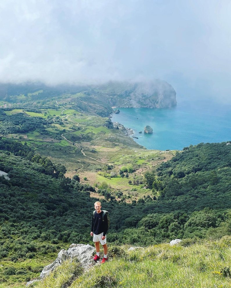 Tim ist zu sehen, von weiter oben auf einem Berg fotografiert und mit einem langen verschlungenen Pfad hinter ihm ins Tal hinunter