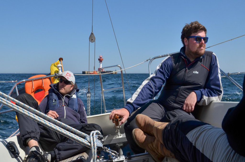 Zwei Männer geniessen die Zeit am Heck eines Bootes. Einer von ihnen hält das Steuer sicher in der Hand.